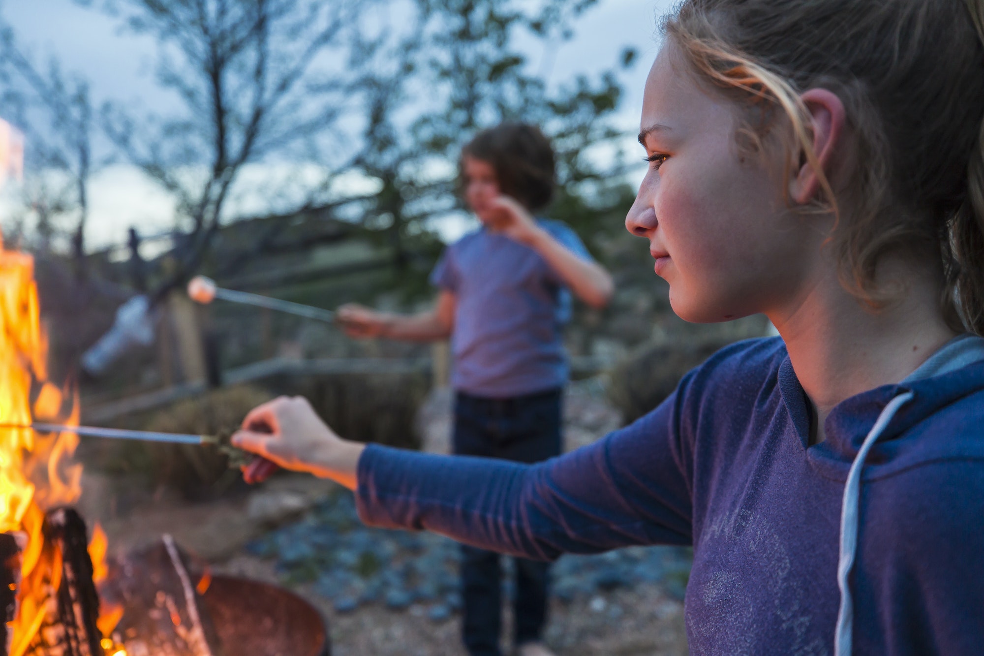A teenage girl making smores with her brother over a fire in a garden at dusk.