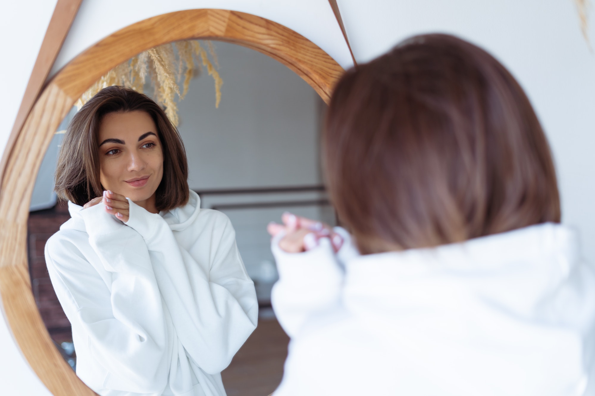 Young woman at home in the bedroom in a warm white hoodie posing in front of the mirror