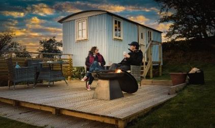 Two individuals sitting around a crackling campfire in front of a shepherd's hut, against a sunset backdrop at Hadrian's Wall campsite