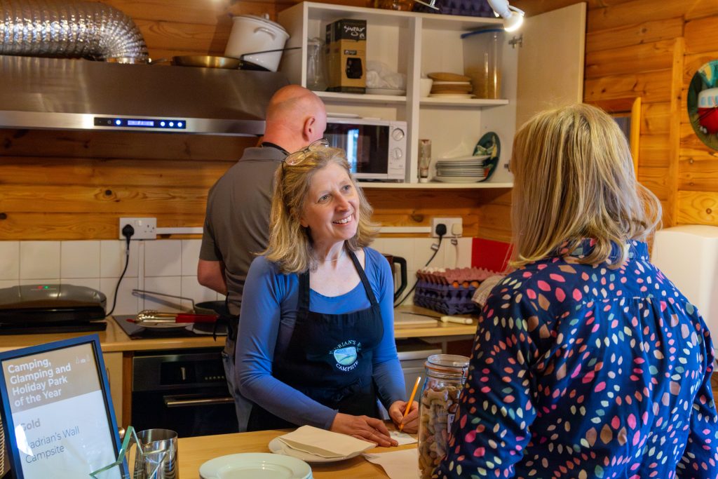 Indoor cafe scene at a the campsite, featuring a friendly woman serving a customer with a warm smile amidst a backdrop of the cafes open kitchen where a man can be seen cooking breakfast sandwhiches.